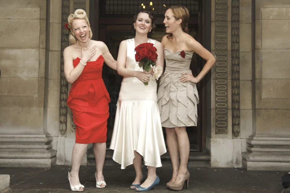 Three women in vintage attire standing in front of a building entrance, sharing a joyful moment. The woman in the middle is holding a bouquet of red roses.