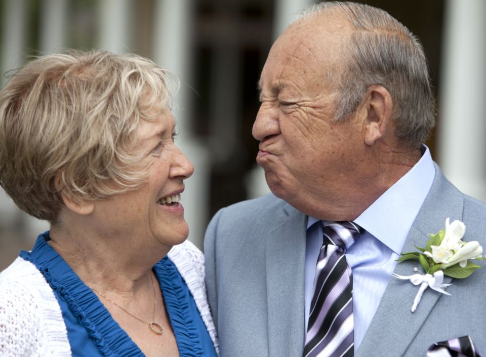 An elderly woman and man dressed in formal attire smiling and looking at each other, possibly at a wedding or special event.
