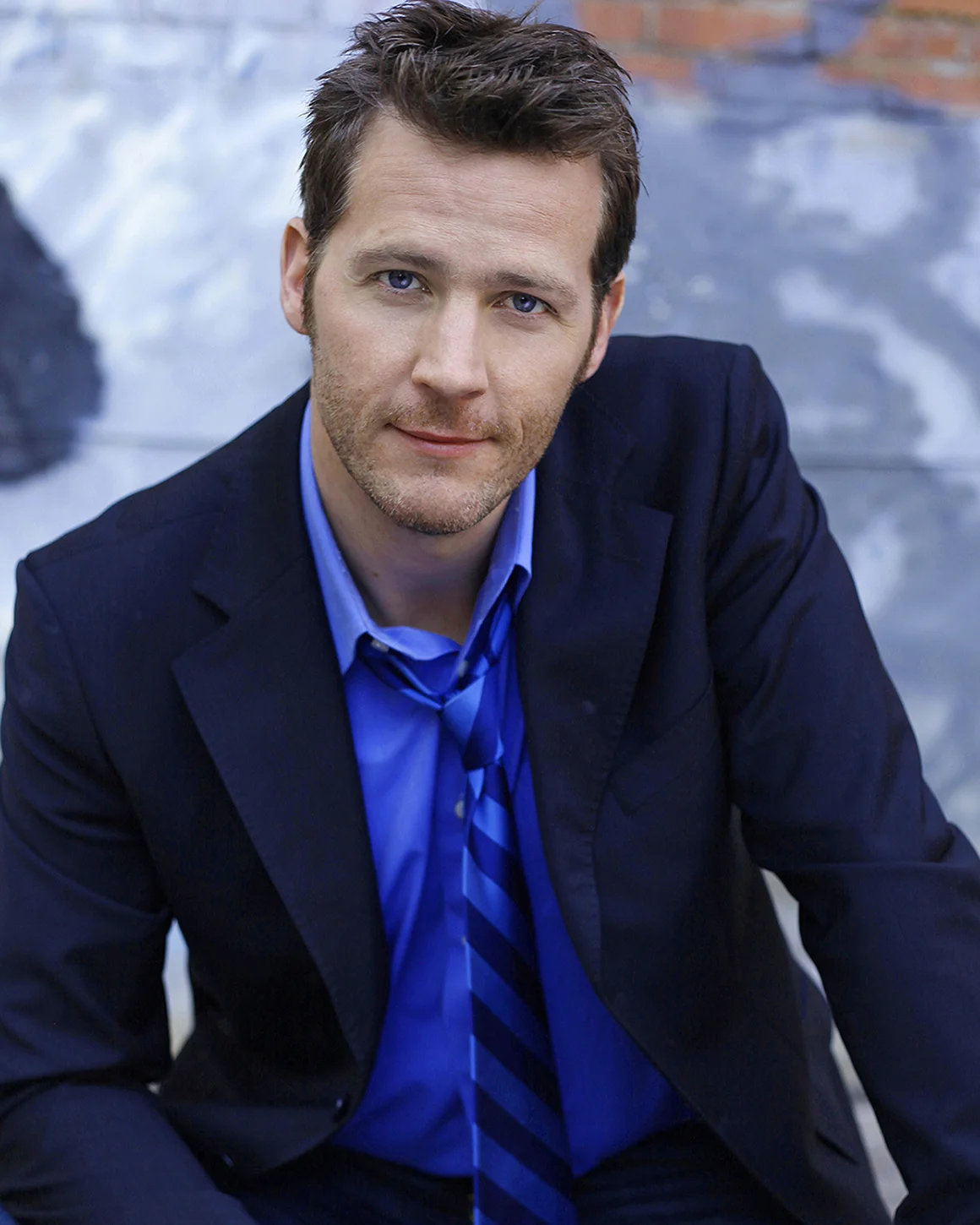A man with short brown hair, blue eyes, and light facial hair wearing a dark suit, blue shirt, and striped blue tie, sitting outdoors against a blurry background of a wall and sky.