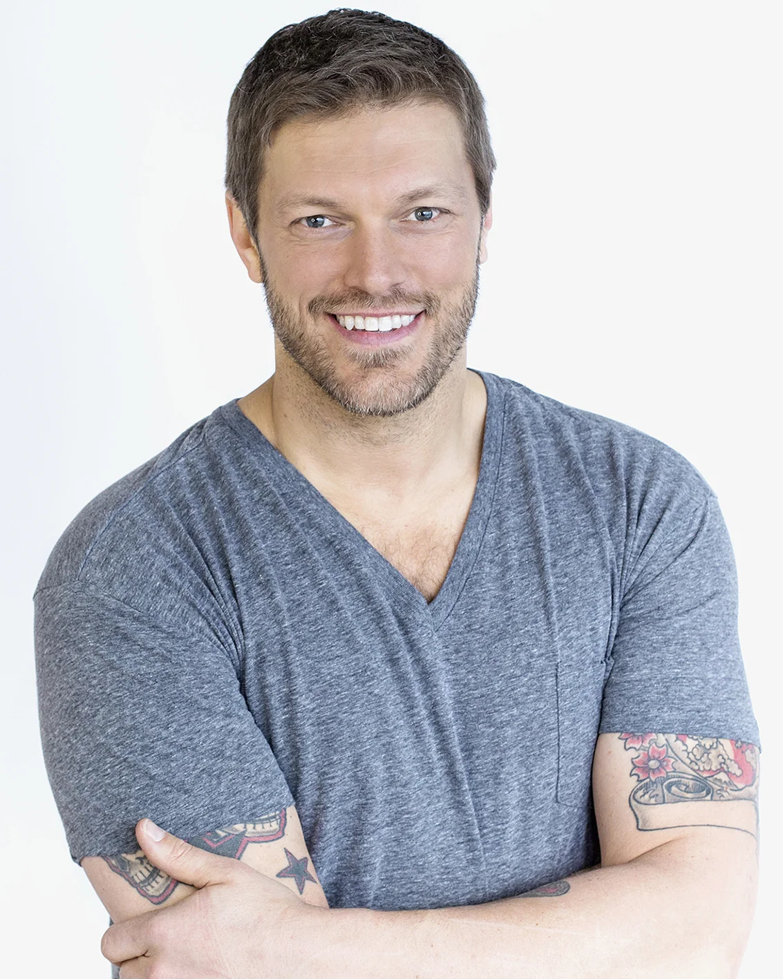 A smiling man with short brown hair, beard, and tattoos on his arms wearing a gray V-neck T-shirt, crossed arms, against a plain white background.