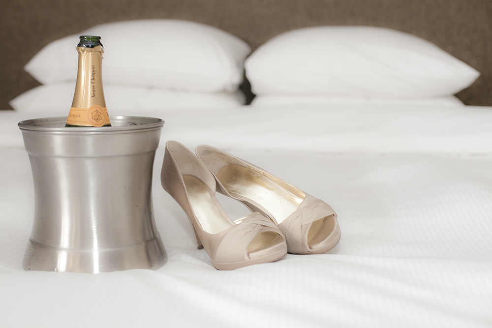 A pair of beige high heels, a bottle of champagne in an ice bucket, and a bed with white pillows.