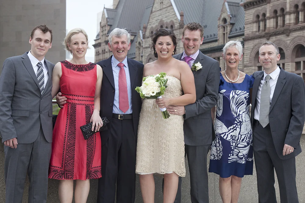 Group of people dressed formally for a wedding, standing outdoors in front of a historic building, smiling, with the bride holding a bouquet of white flowers.