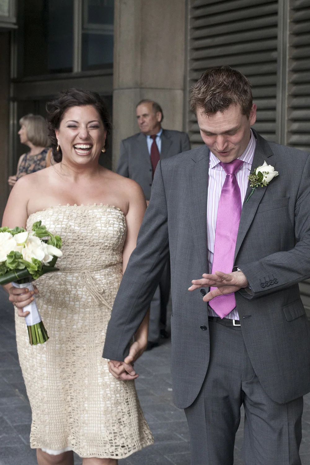 A woman in a strapless dress laughing and holding a bouquet of white flowers, standing next to a man in a gray suit with a pink tie, who is looking at his wristwatch, holding hands with each other during a wedding celebration.