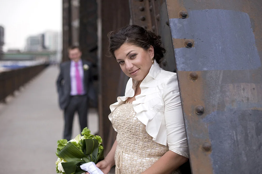 A woman in a wedding dress holding a bouquet, leaning against a rusted metal structure, with a man in a suit standing in the background on an outdoor urban walkway.