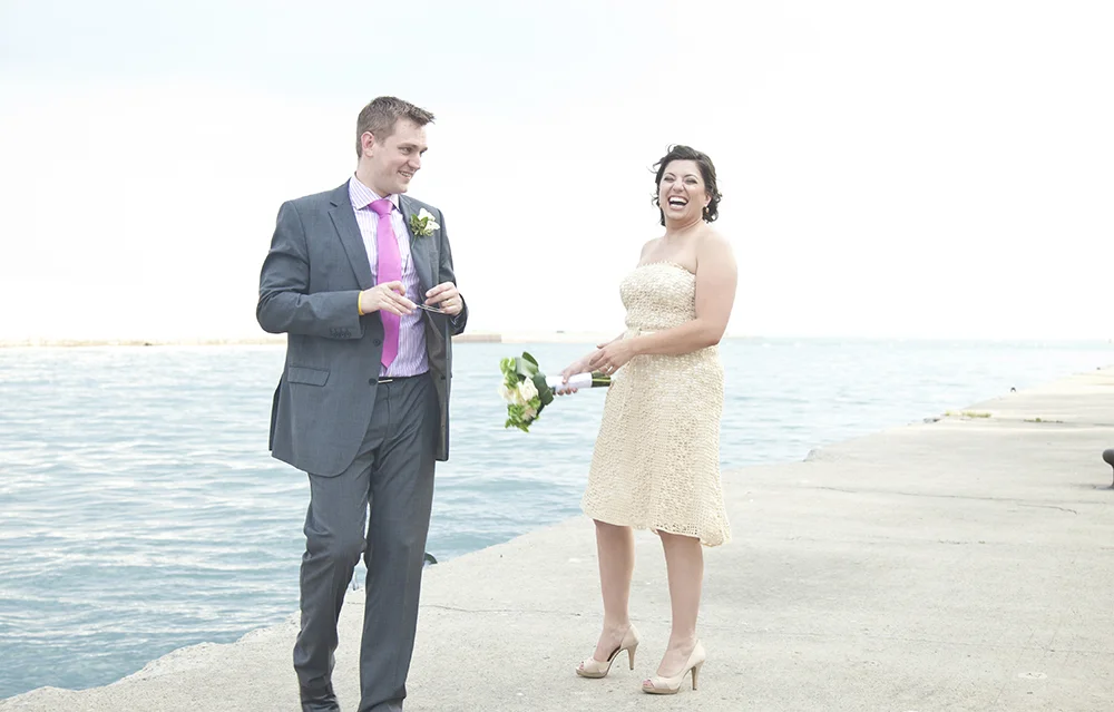 A couple in wedding attire standing on a pier by the water, smiling and laughing during their wedding photoshoot.
