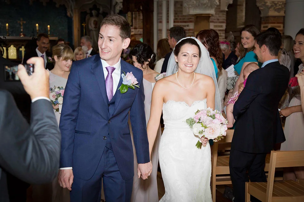 A bride and groom holding hands and smiling during their wedding ceremony in a church, surrounded by family and friends.