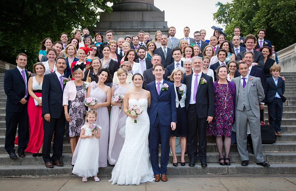 A large group of people gathered on outdoor steps for a wedding photo, with a bride and groom in the center. The bride is in a white wedding dress holding a bouquet, and the groom is in a navy suit with a purple tie. The group includes men, women, an