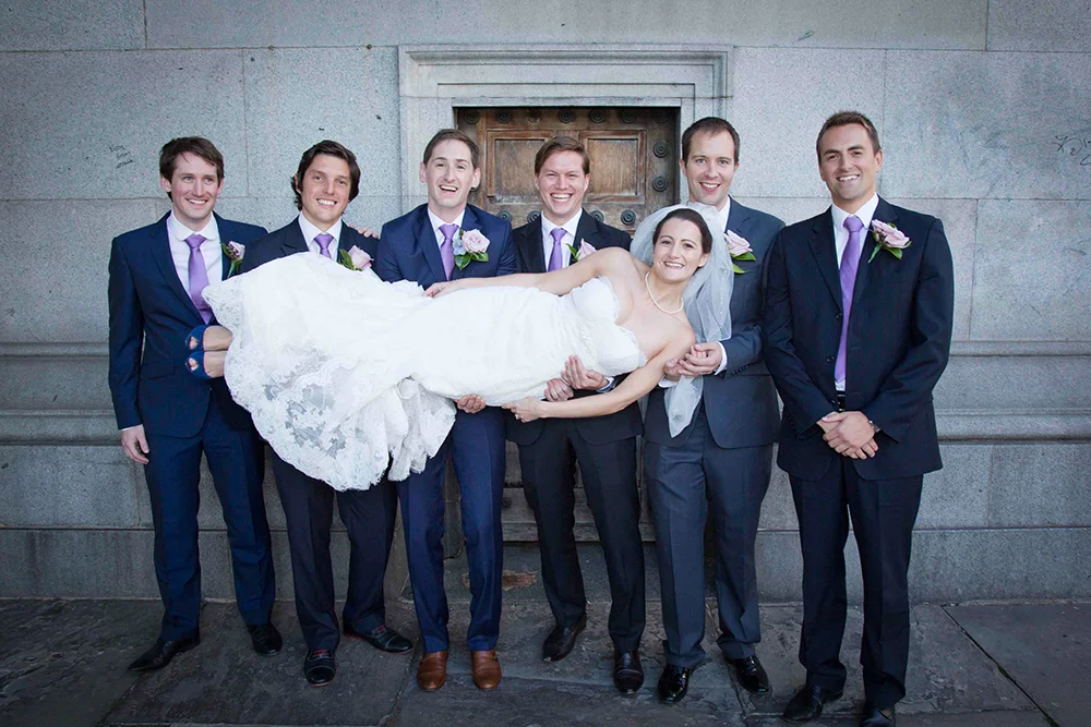 Group of men in suits holding a bride horizontally in front of a stone wall.