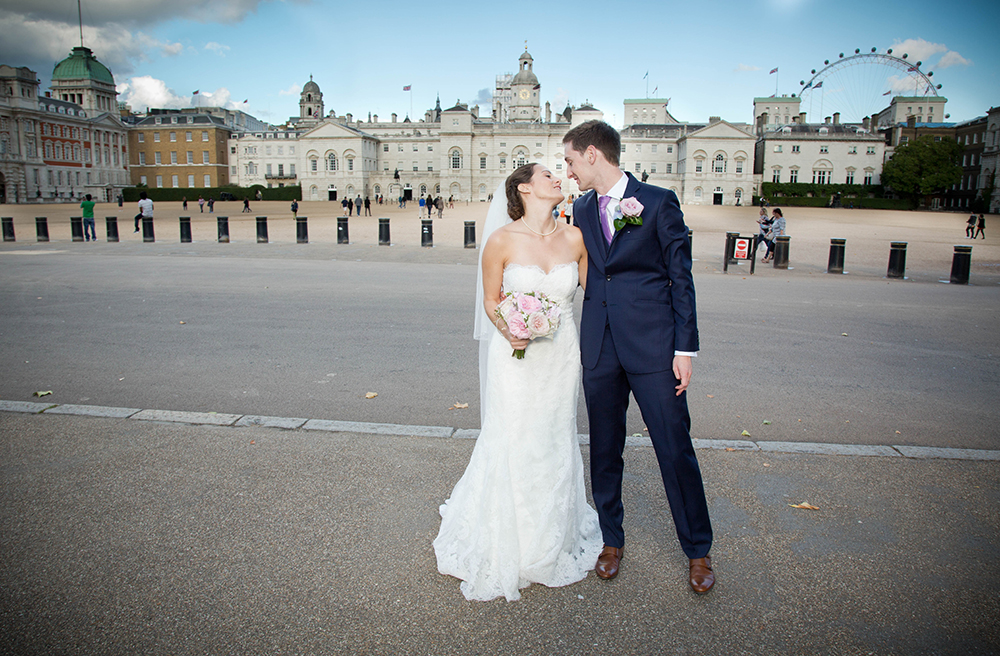 A bride in a white wedding dress holding a bouquet, and a groom in a navy suit with a boutonniere, sharing a kiss outside an historic building with a Ferris wheel in the background.