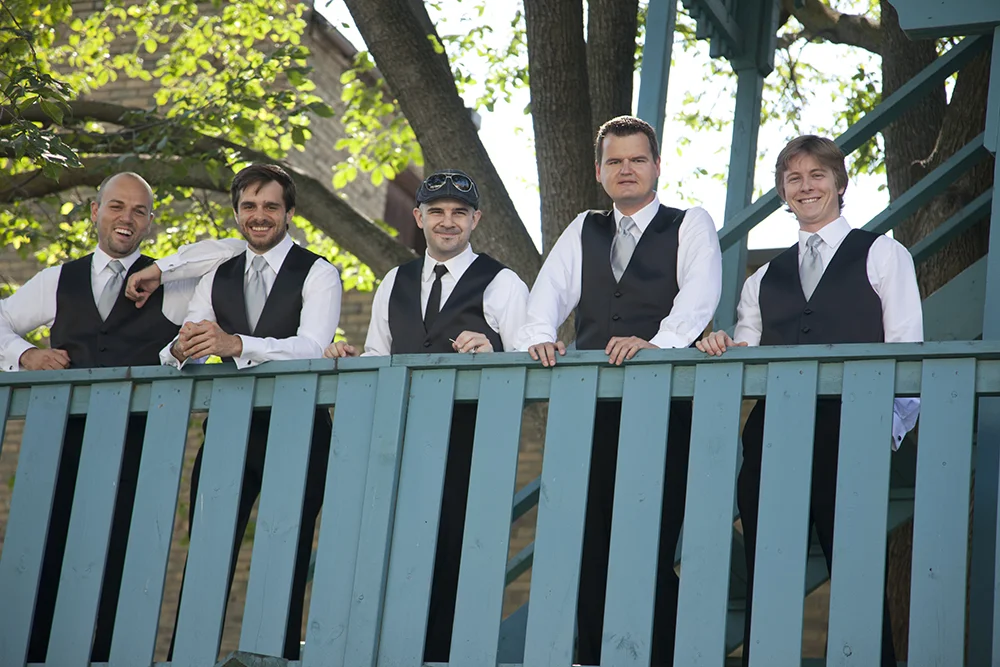 Five men in formal black vests and white shirts stand behind a blue railing outdoors near a large tree, smiling for the photo.