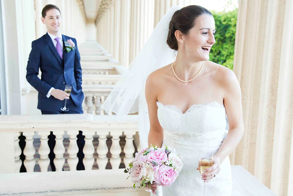 Bride in a white strapless wedding dress holding a bouquet of pink roses and glasses of champagne, smiling and looking to the side, with a groom in a dark suit and purple tie in the background.