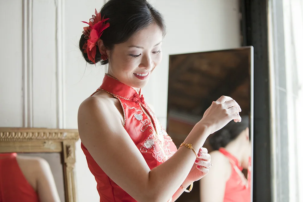 Young woman in a red dress with floral embroidery, smiling while adjusting her bracelet in front of a mirror, with a window letting in natural light.