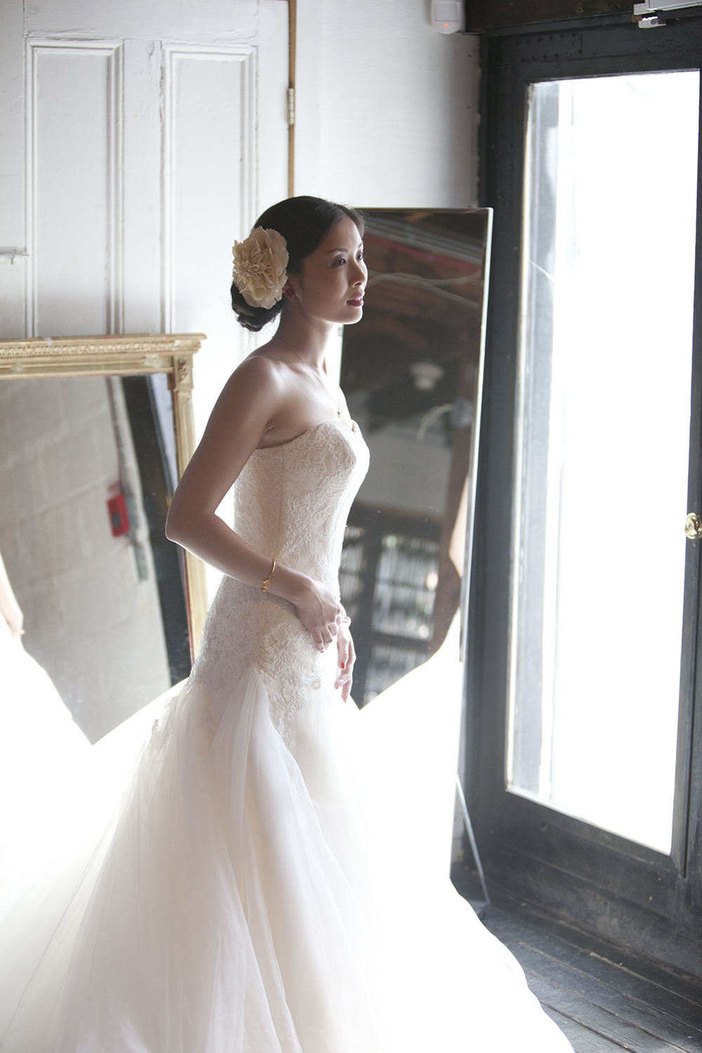 A woman in a strapless wedding dress with lace details and a tulle skirt, standing by a glass door, facing sideways, with a large flower hair accessory.