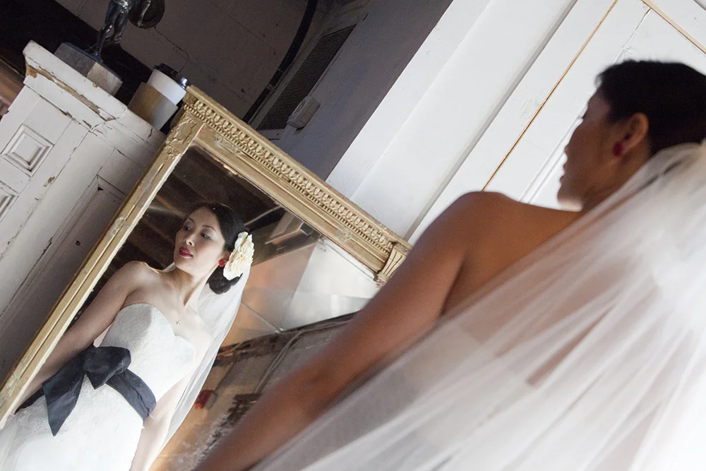 A bride in a white wedding dress with a black bow, looking into a mirror.
