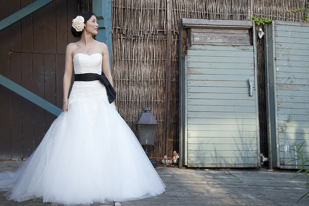 A woman in a white strapless wedding dress with a black sash and large white flower hair accessory standing outdoors.