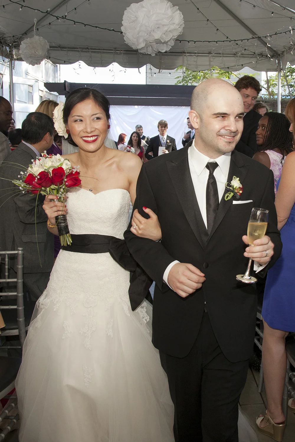 Bride in a strapless white wedding dress holding a bouquet of red and white flowers, smiling, walking arm-in-arm with groom in a black tuxedo and holding a glass of champagne at a wedding reception.