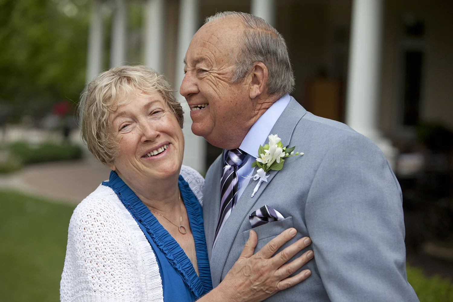 An elderly couple smiling and embracing outdoors, dressed in formal attire with a boutonniere on the man's suit lapel, in front of a house.