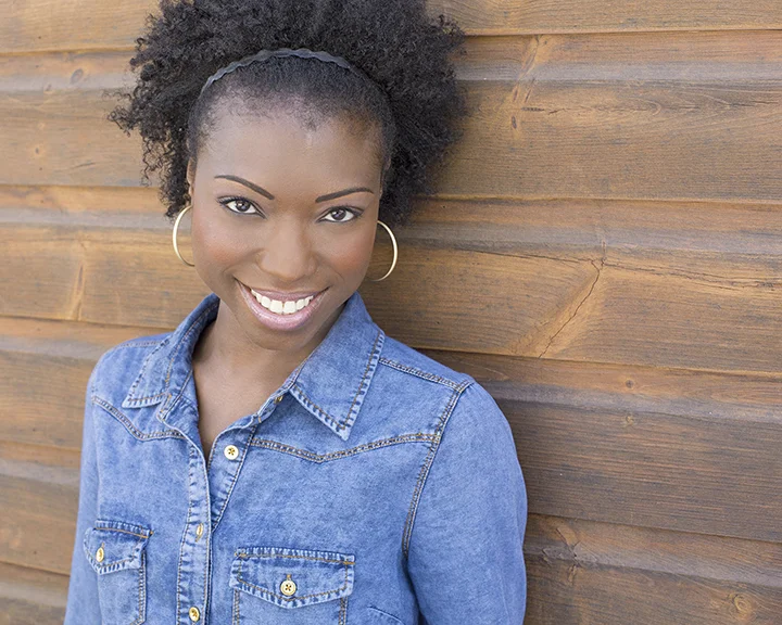 A smiling woman with natural curly hair wearing a denim jacket and hoop earrings, standing against a wooden wall.