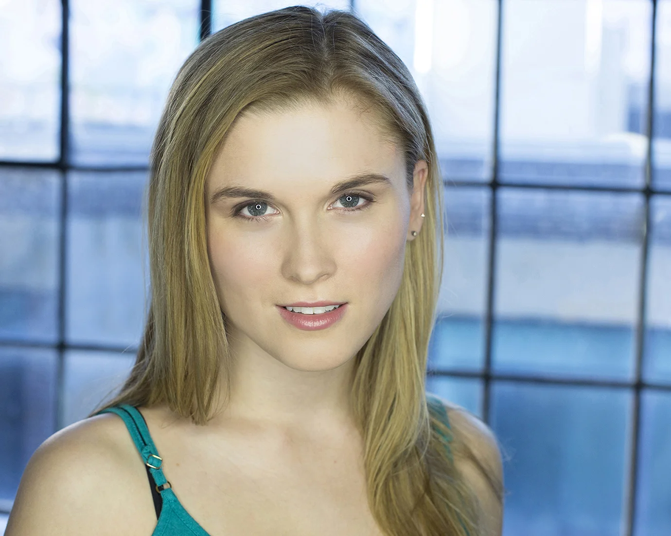 A young woman with long blonde hair, blue eyes, and fair skin, smiling slightly, wearing a teal tank top, in front of a large window with a grid pattern, during daytime.