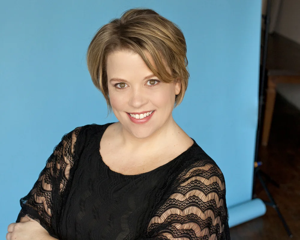 A woman with short, light brown hair smiling, wearing a black lace top, in front of a blue background.