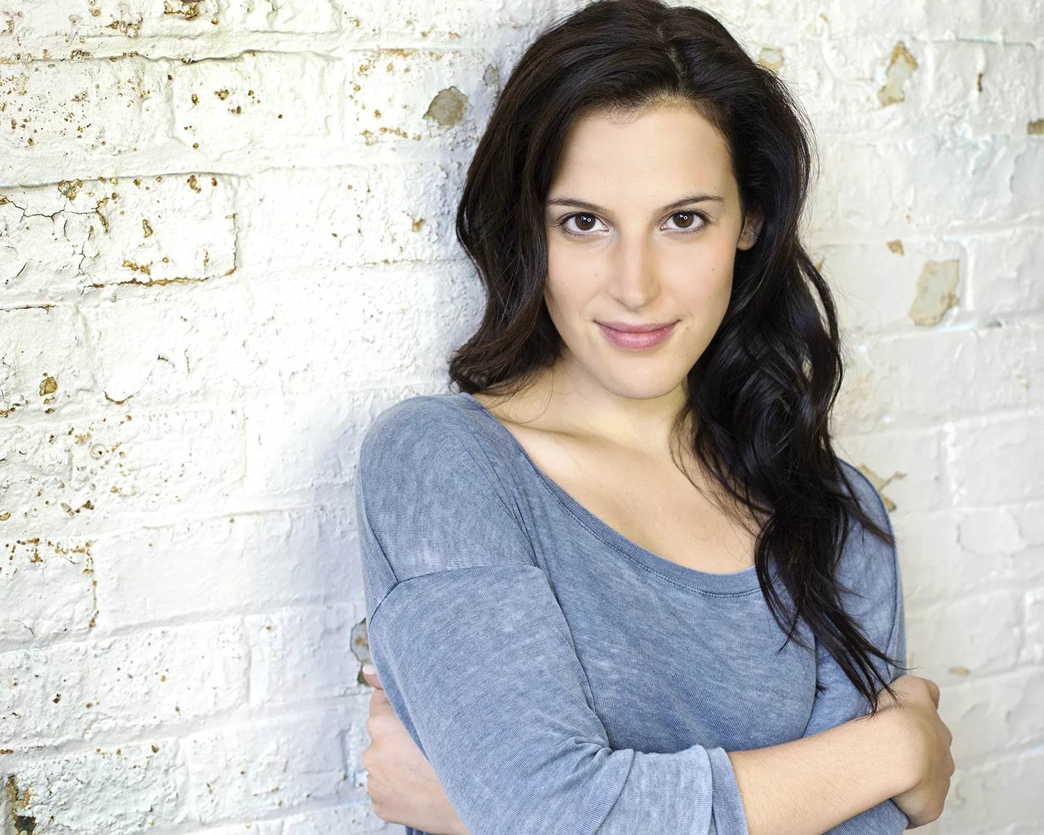 A woman with dark, wavy hair wearing a blue top, standing against a white brick wall, looking at the camera with a slight smile.