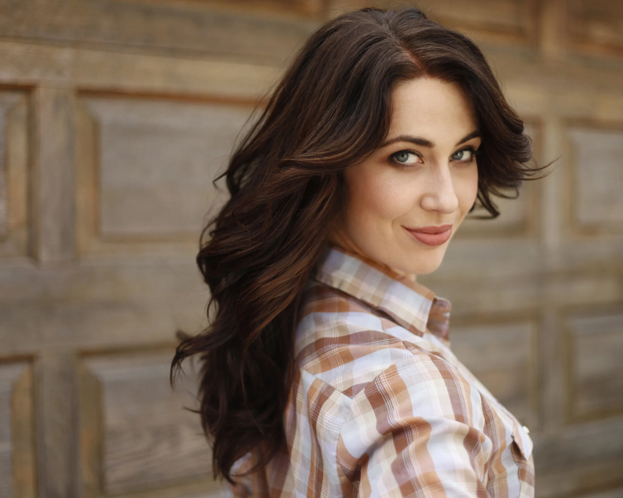 A woman with long, wavy brown hair and piercing blue eyes looking at the camera and smiling, wearing a plaid shirt in front of a wooden background.
