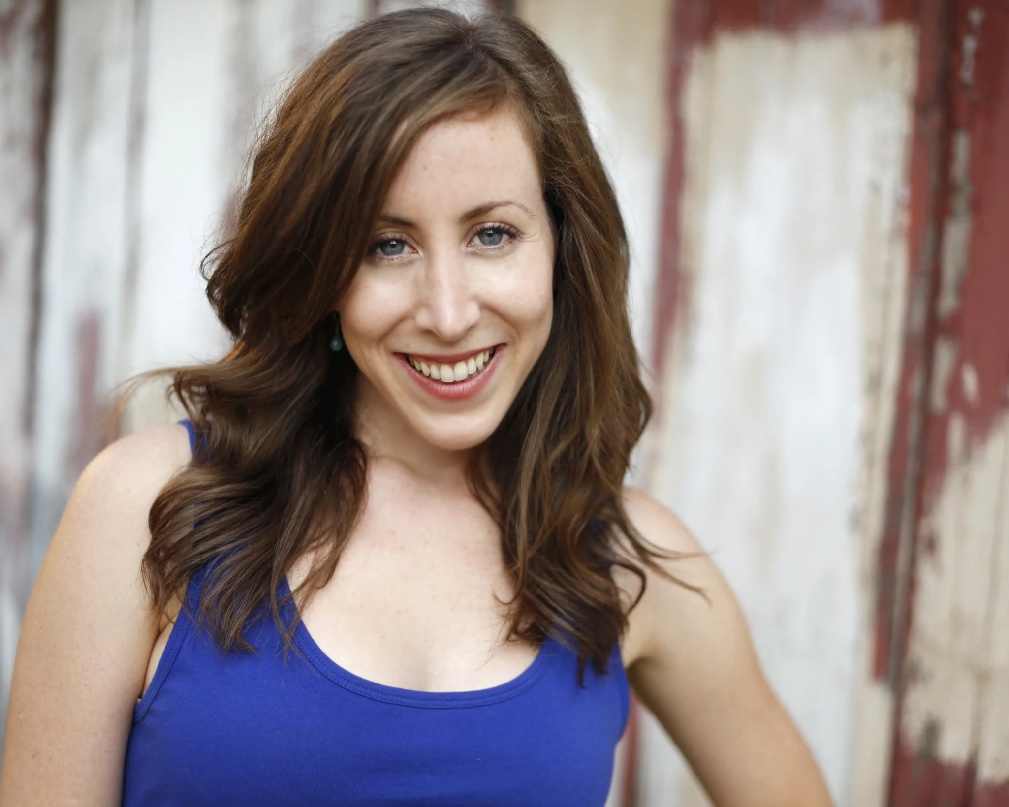 A woman with long, wavy brown hair, smiling and wearing a blue sleeveless top, standing against a rustic wooden wall.