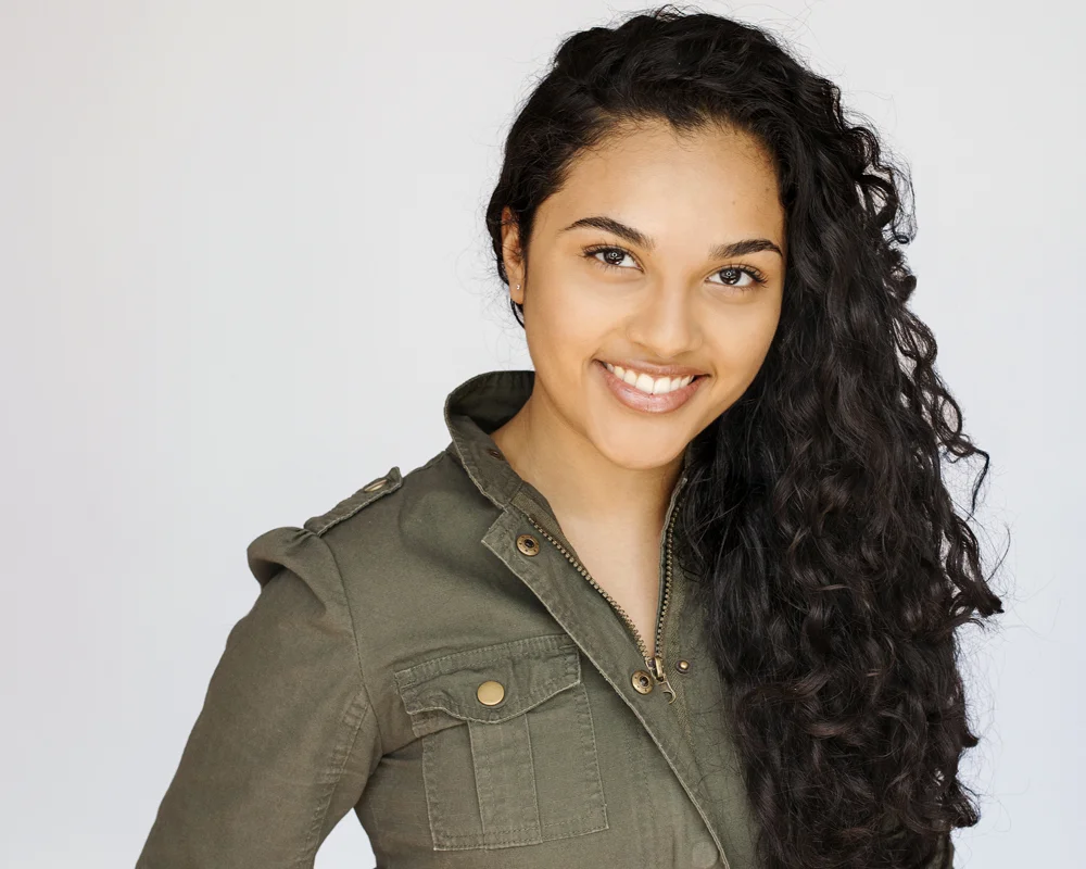 Portrait of a young woman with long, curly black hair smiling at the camera, wearing an olive-green jacket.