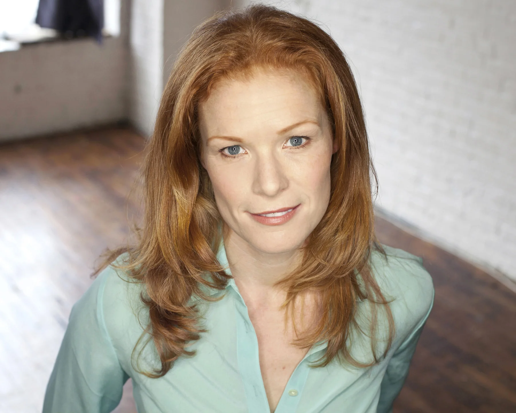 Portrait of a woman with red hair and blue eyes, wearing a light blue shirt, looking at the camera, in a bright room with a hardwood floor and white brick wall.
