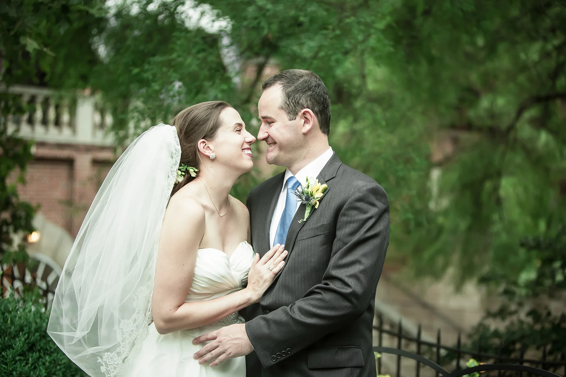 A bride and groom on their wedding day outdoors, smiling and holding each other close, with green trees and a brick building in the background.
