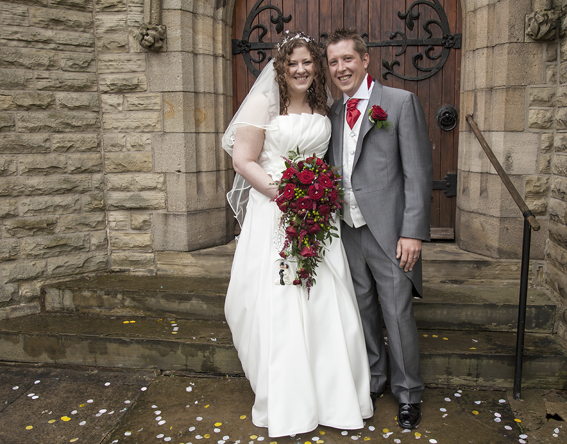 A bride and groom happily standing in front of a stone and wood door, dressed in wedding attire, with confetti on the ground.