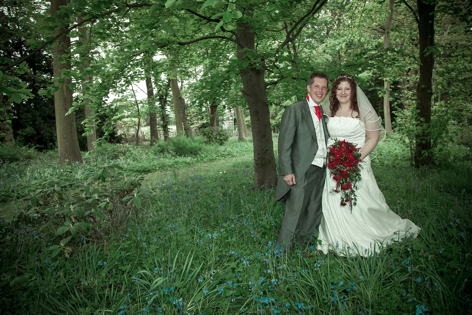 A newlywed couple in wedding attire stands together in a lush green forest. The bride holds a large bouquet of red flowers, and the groom wears a gray suit with a red tie. They are smiling and appear happy.