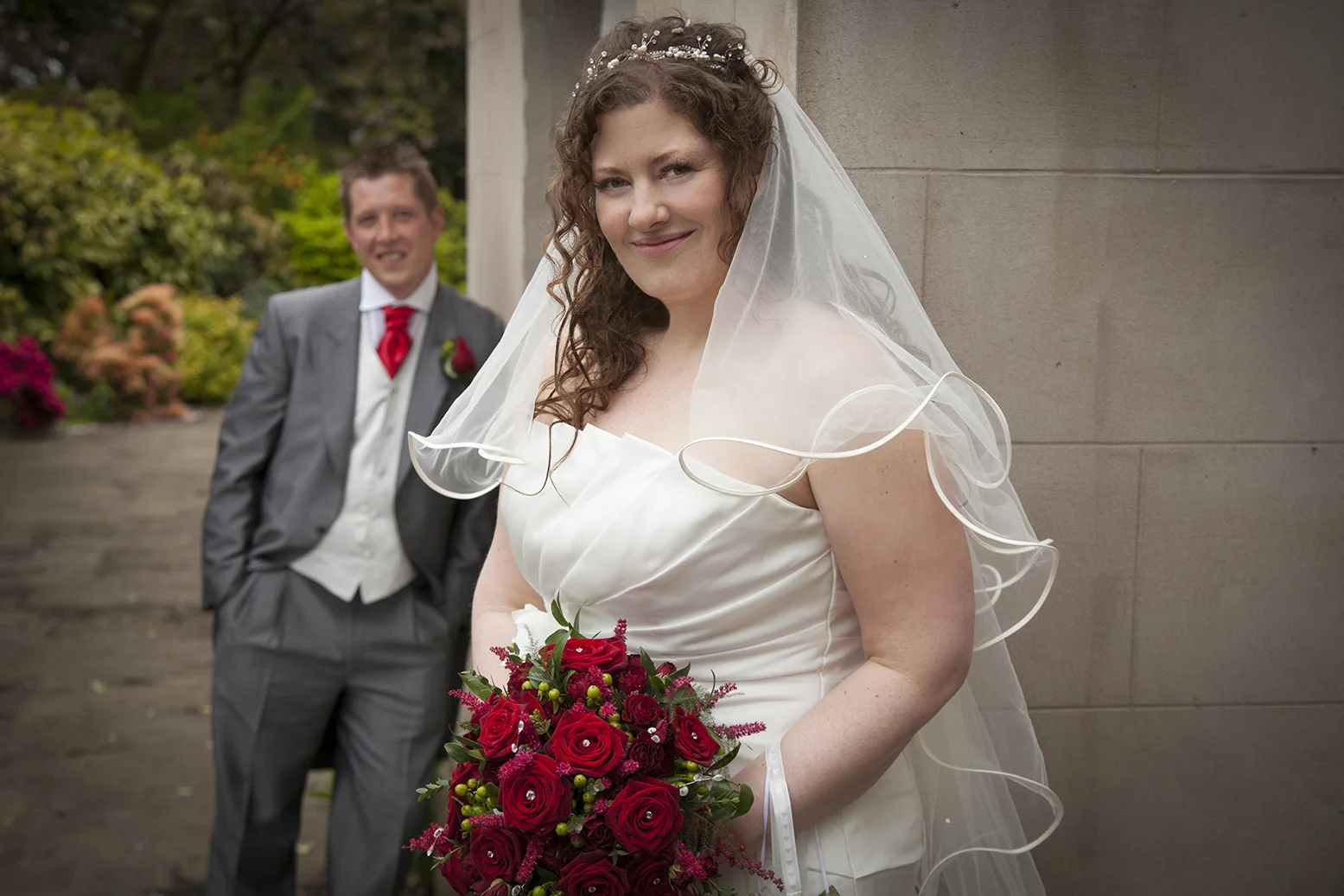 A bride with curly hair holding a bouquet of red roses, wearing a white wedding dress and veil, standing outdoors with a groom smiling in the background, dressed in a gray suit and red tie.