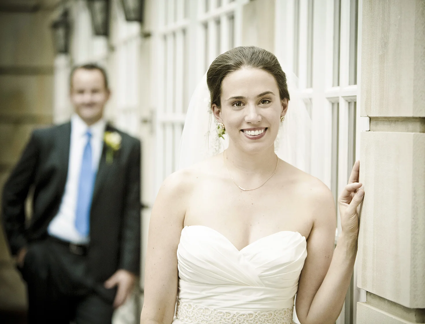 A smiling bride in a white strapless wedding dress posing near a window with her hand on the wall, while a groom in a suit with a blue tie stands in the background.