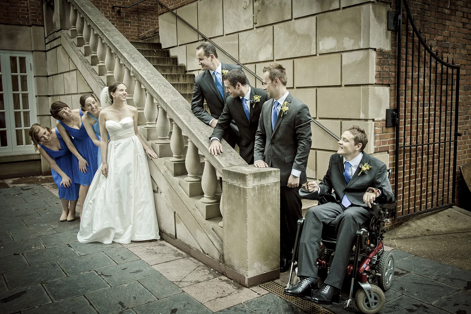 A bride in a white wedding dress stands beside five bridesmaids in matching blue dresses, laughing and leaning forward. Five groomsmen in suits with blue ties and boutonnières lean over a stone balustrade, smiling and engaging with the bride and brid