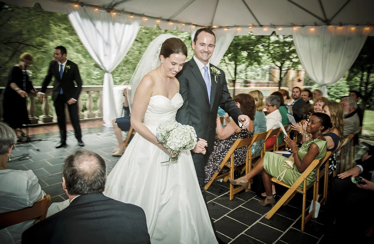 A bride and groom walking down the aisle during their wedding celebration, with guests seated on wooden chairs under a white canopy with draped curtains and string lights in an outdoor setting surrounded by trees.