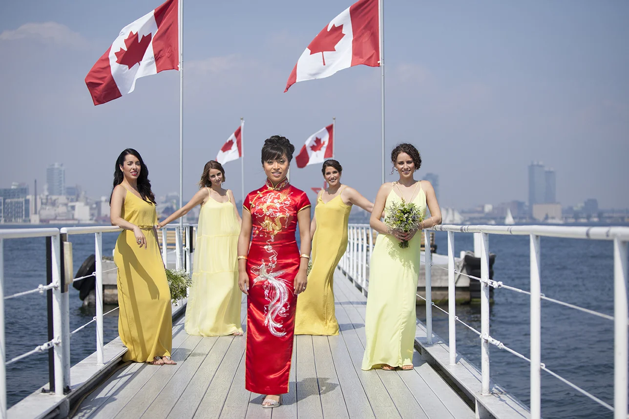 Women in colorful dresses on a boat dock, with Canadian flags on poles in the background, city skyline visible across the water.