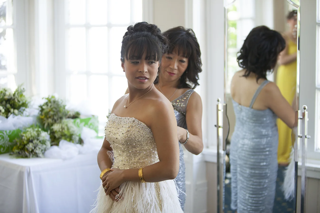 Women getting ready, one in a gold and white dress with feather details, another helping her with her dress in a bright room with flowers on a table.