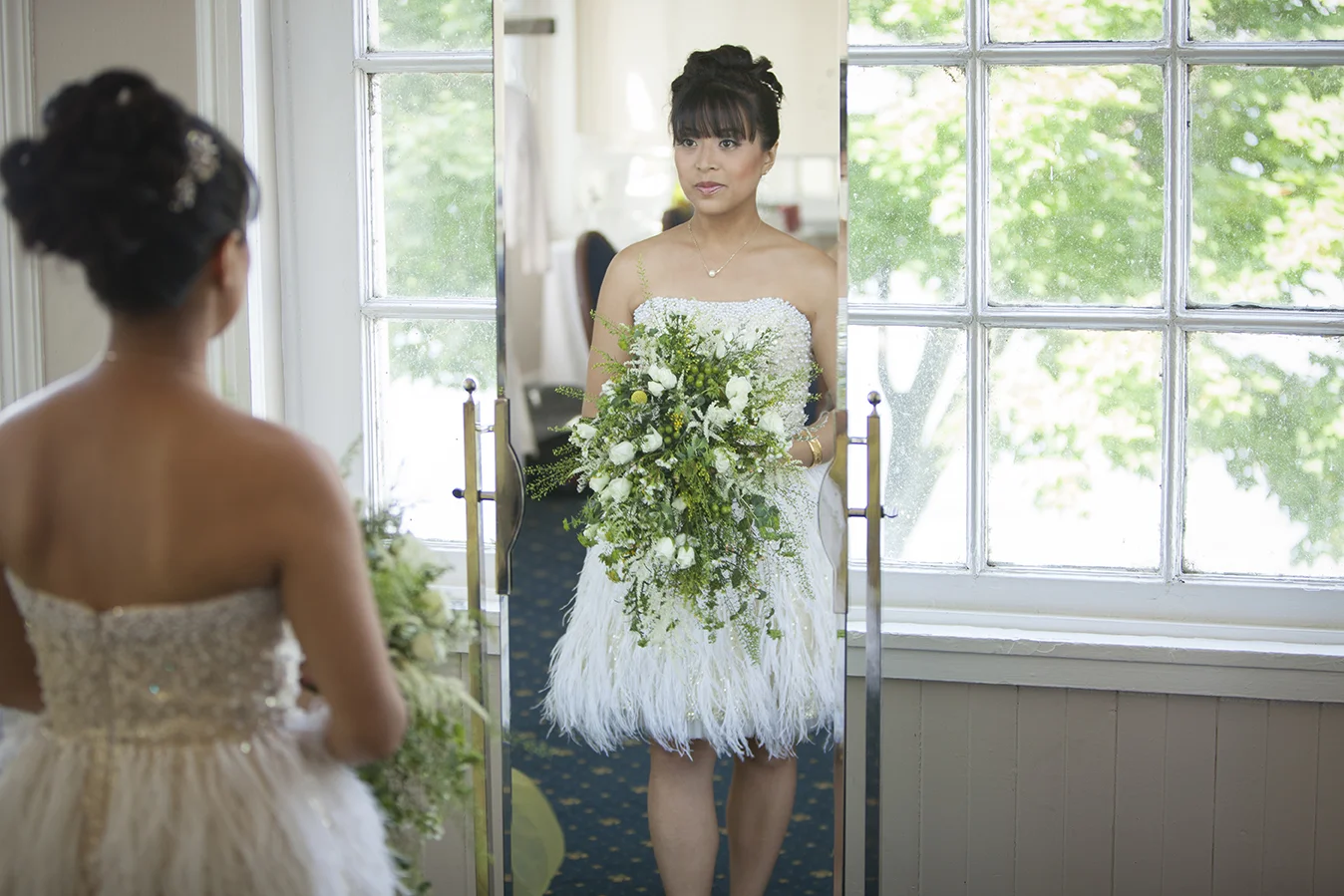 Two women in wedding dresses, one facing a mirror and the other with her back to the camera, indoors near a window.