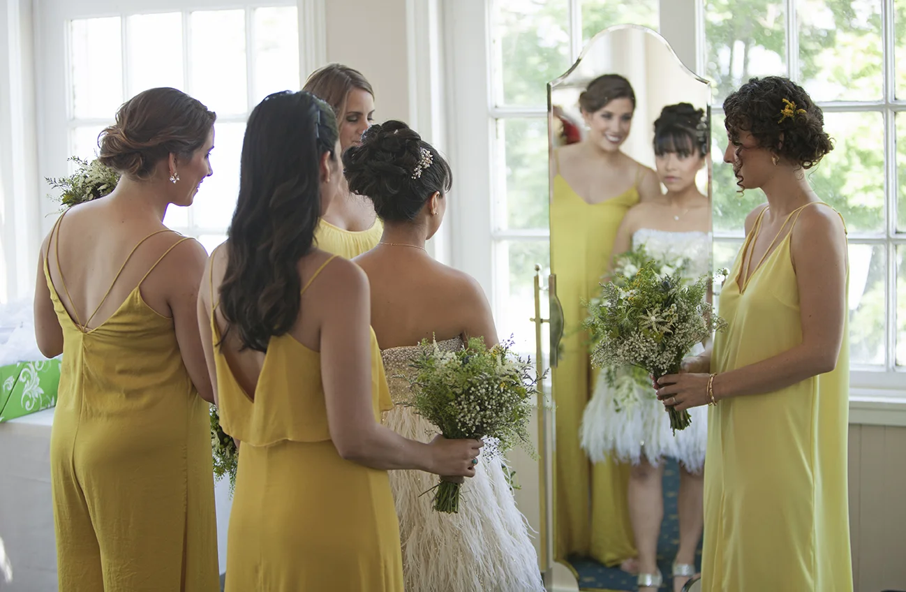 Bridesmaids in yellow dresses giving a bride a bouquet in front of a mirror.