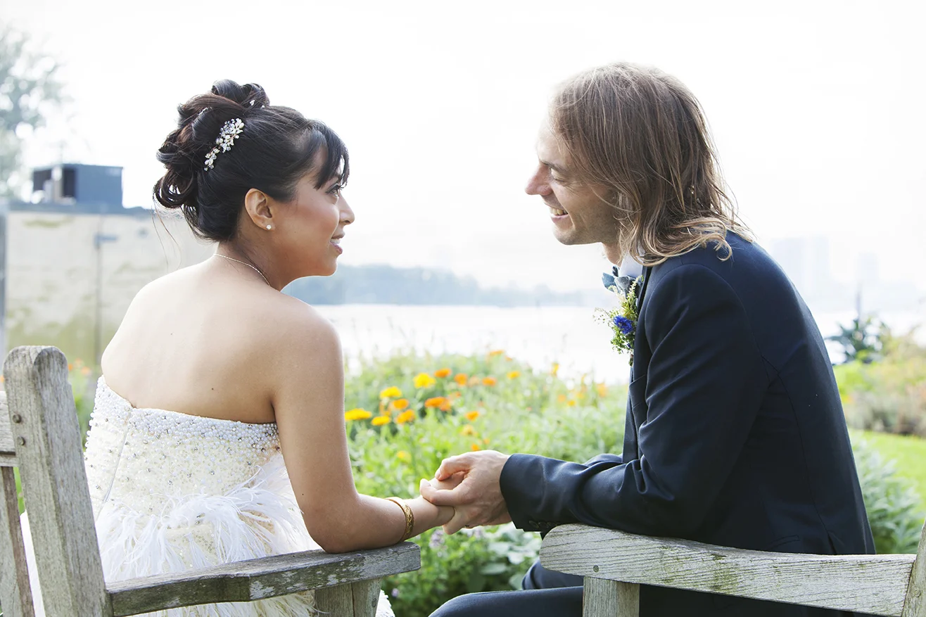 A bride and groom sitting on a park bench, holding hands and looking at each other, outdoors with flowers and a body of water in the background on a bright day.