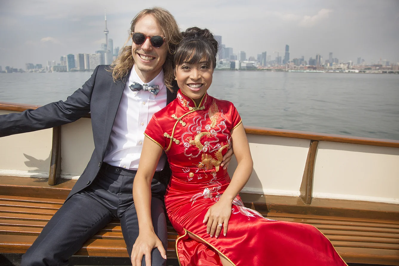 A man and woman smiling and sitting on a boat with Toronto skyline in the background. The woman is wearing a traditional red Chinese dress with gold embroidery, and the man is wearing a dark suit with a bow tie and sunglasses.