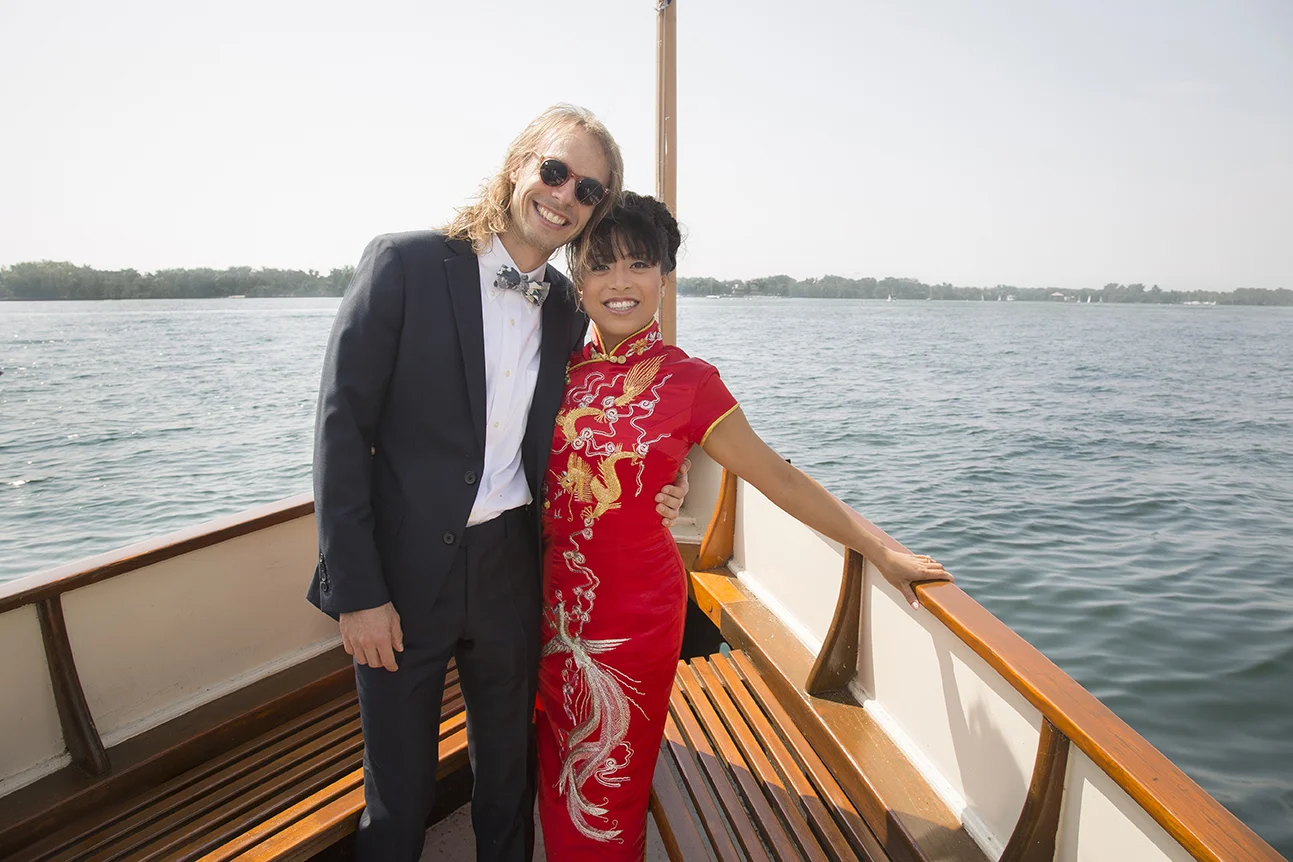Two smiling people, a man in a dark suit with sunglasses and a woman in a red traditional dress, stand on a boat with water and a distant shoreline in the background.