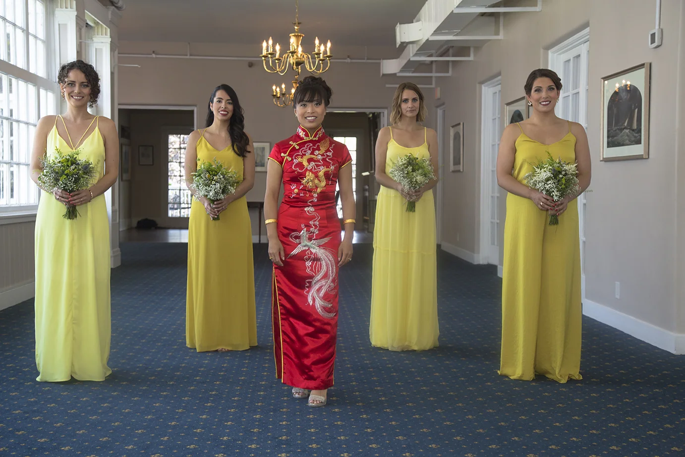 Bride in a red traditional Chinese wedding dress with gold embroidery, surrounded by four bridesmaids in yellow dresses holding bouquets in an indoor setting with chandeliers and framed pictures on the walls.
