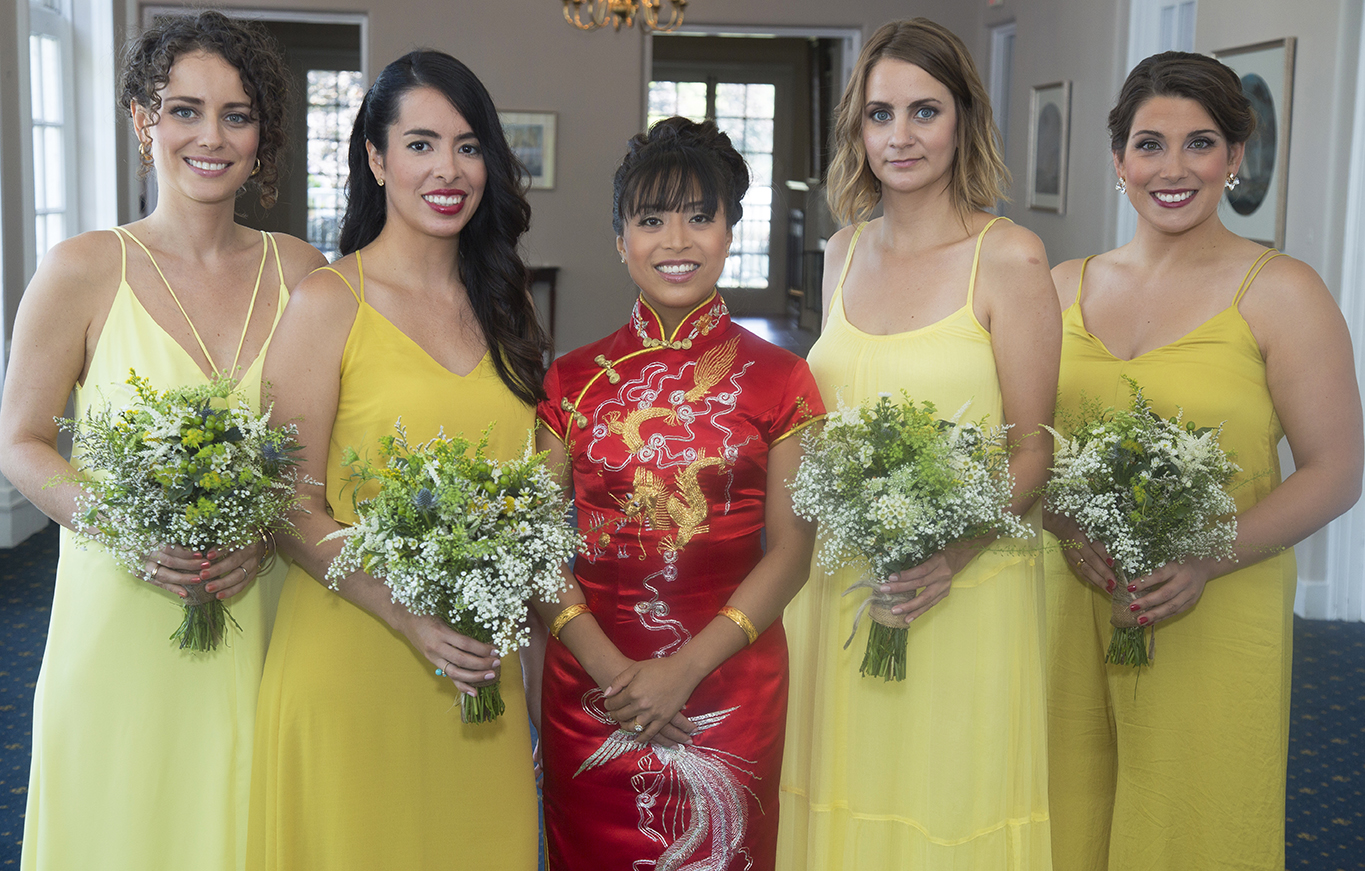 Five women standing indoors, four wearing yellow dresses and holding flower bouquets, and one wearing a red traditional Chinese dress with gold embroidery, smiling at the camera.