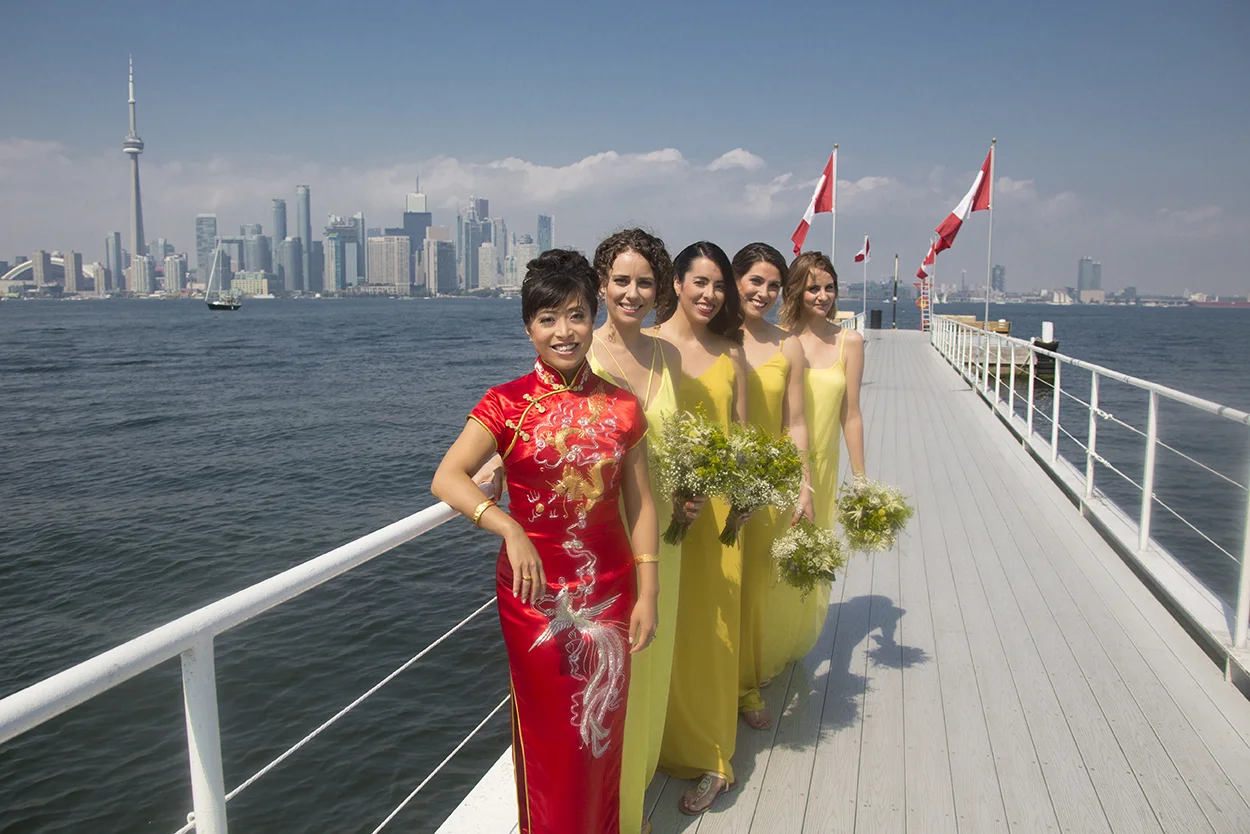 Group of five women standing on a dock with a city skyline in the background, four women in yellow dresses holding bouquets, one woman in red traditional Chinese dress.