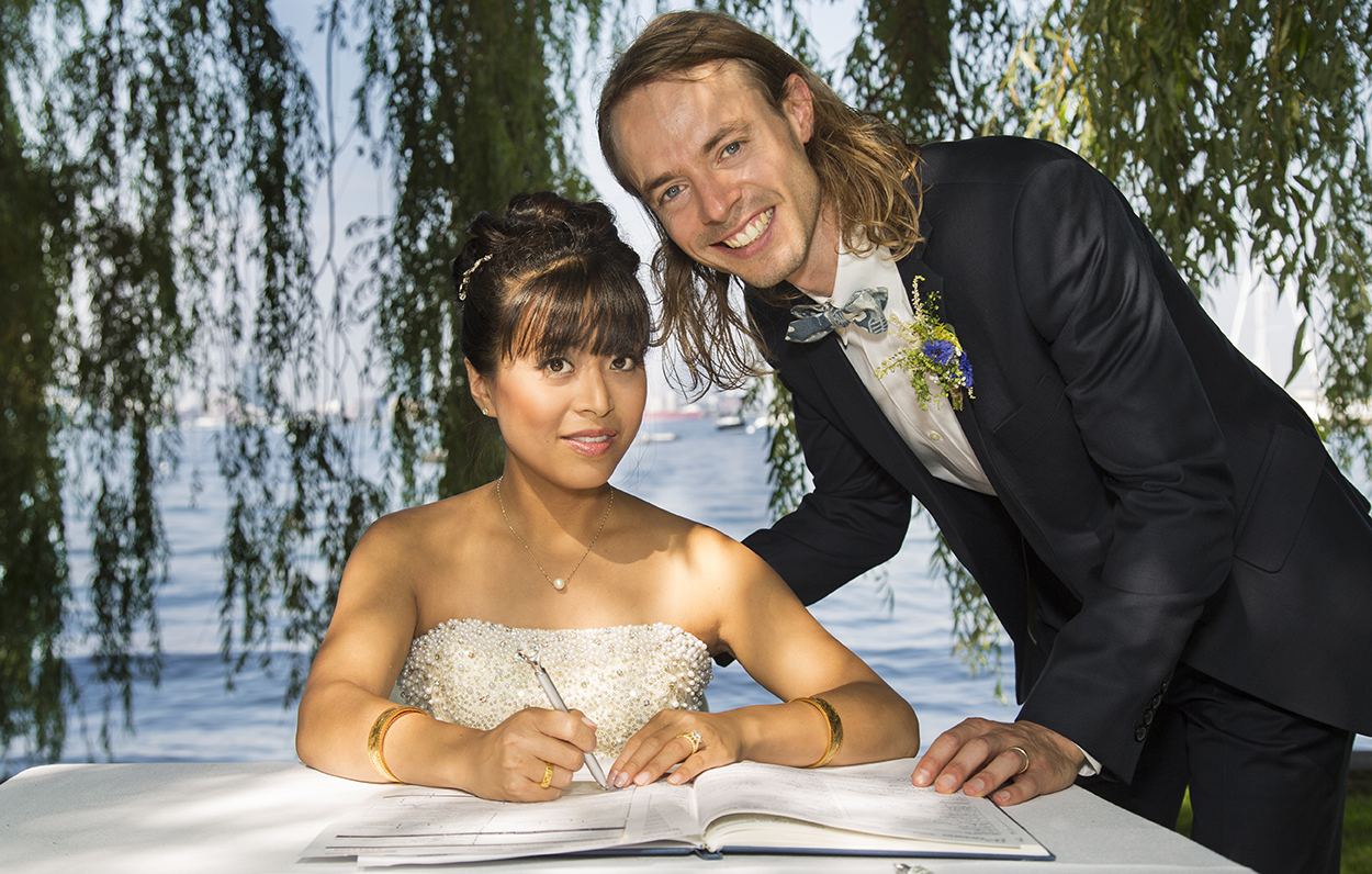 A bride signing a document at a table outdoors near a lake, with a groom standing beside her and leaning over, both smiling.