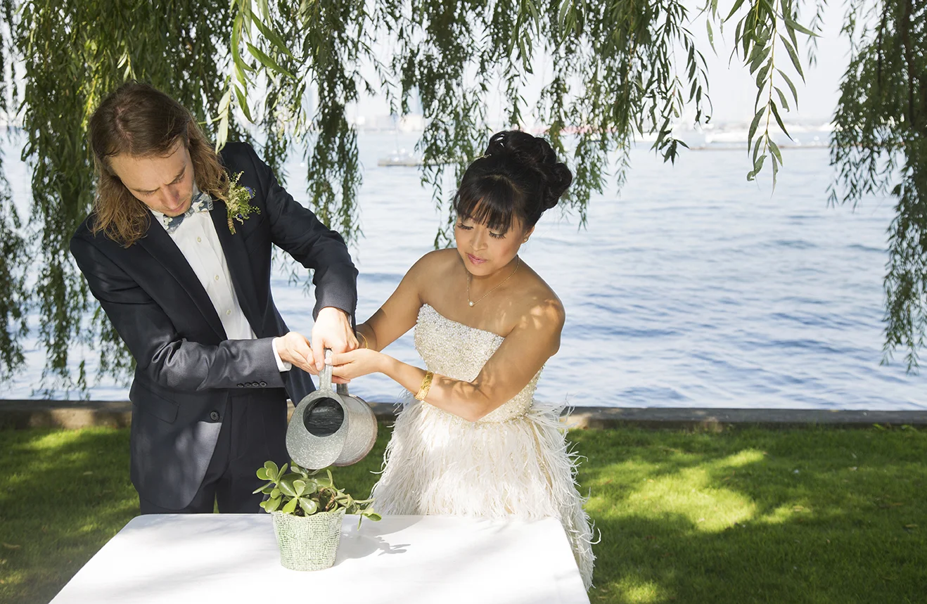 A couple dressed in wedding attire watering a potted plant at a lakeside outdoor wedding ceremony.