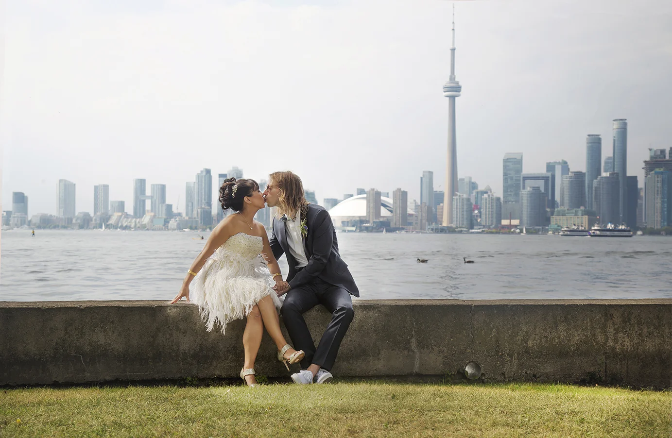 Bride and groom kiss beside a body of water with a city skyline, including the CN Tower, in the background.