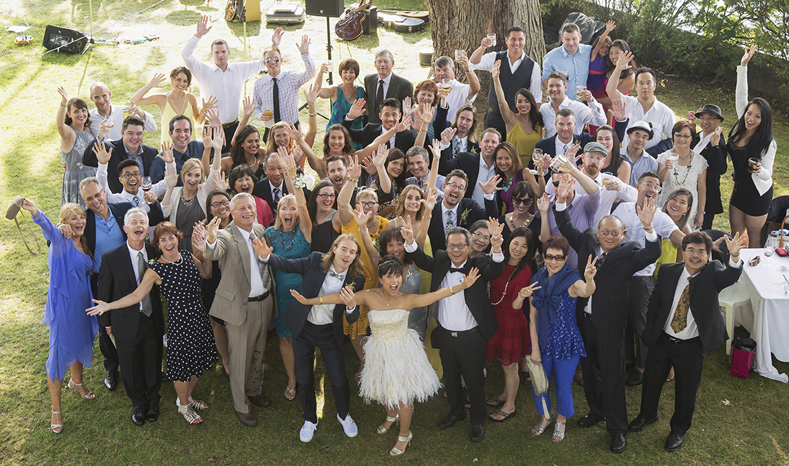 A large group of people in formal and casual attire celebrating outdoors, waving, and smiling, with a table set up on the right and musical instruments in the background.